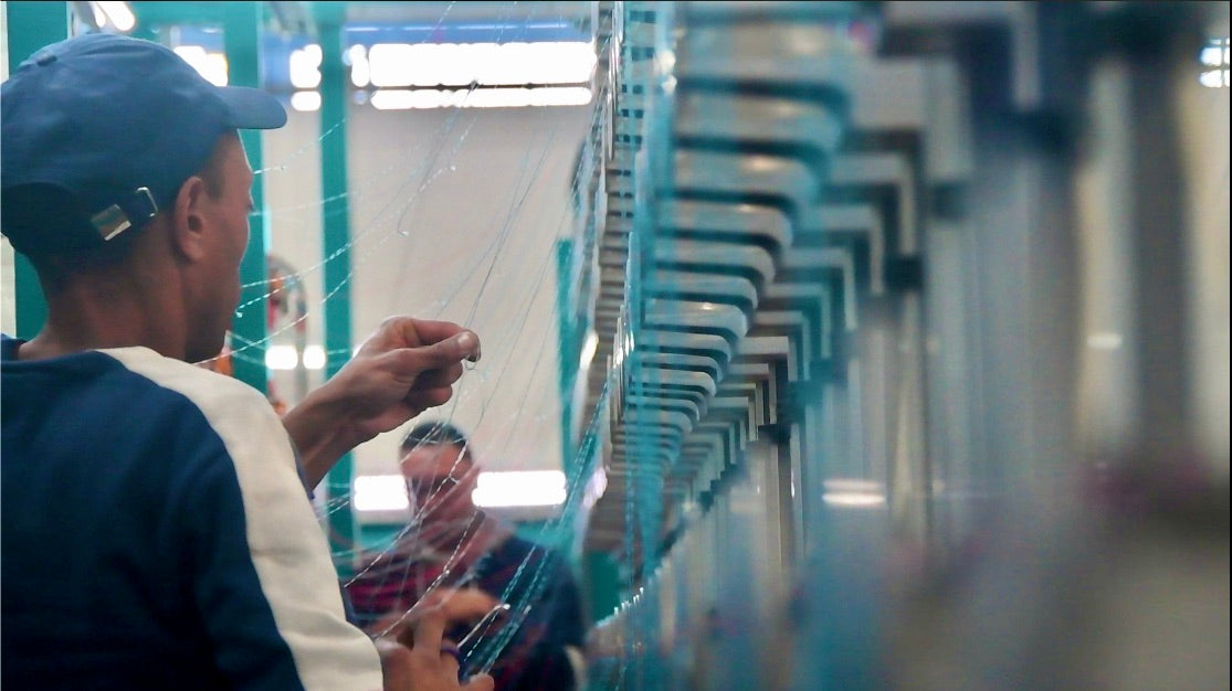 Worker manually threading yarns on an industrial textile weaving machine inside a factory.